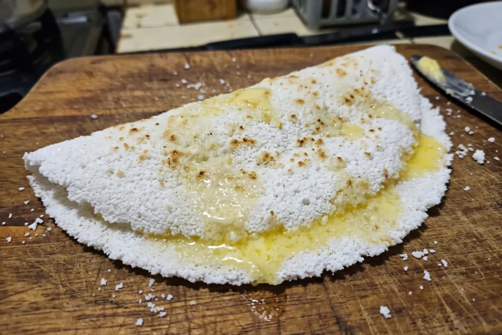 Tapioca com manteiga dobrada ao meio saindo fumaça em um prato branco sobre mesa de madeira